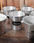 Five Go Home Vintage Champagne Buckets, upcycled and featuring unique logos and inscriptions, are arranged on a wooden surface with warm wooden paneling in the background.