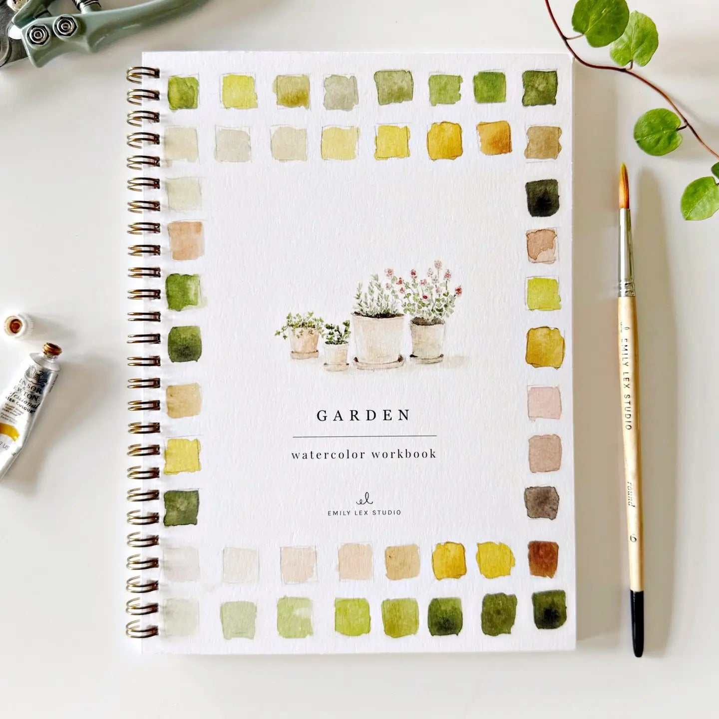Faire's beginner-friendly "Watercolor Workbook" has a charming spiral binding and features painted plant pots on the cover. The title is surrounded by delicate watercolor patches in green, yellow, and brown, while a paintbrush and watercolor tubes rest gracefully nearby.