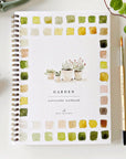 Faire's beginner-friendly "Watercolor Workbook" has a charming spiral binding and features painted plant pots on the cover. The title is surrounded by delicate watercolor patches in green, yellow, and brown, while a paintbrush and watercolor tubes rest gracefully nearby.