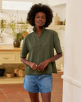 A woman with curly hair wears the Frank & Eileen Patty Popover SS Henley in olive green and denim shorts, standing in a bright kitchen with plants and baskets in the background.