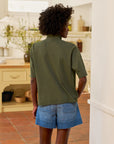 A person with short curly hair, wearing a Frank & Eileen Patty Popover SS Henley in olive green and denim shorts, stands in a bright kitchen with tile floors and light-colored cabinets, facing away from the camera.
