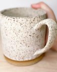A hand holds Faire’s Large Speckle Mug, an 18oz hand-thrown ceramic with a curved handle, a textured off-white surface with brown speckles, and a brown base, placed on a light wooden surface.