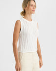 A woman with blonde hair in a loose bun wears the Carrick Tank by One Grey Day, a relaxed fit, sleeveless white knit top, paired with light beige pants. She stands against a plain white background and gazes slightly to the side.