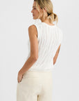 A woman with blonde hair in a low ponytail wears the One Grey Day Carrick Tank, a relaxed fit sleeveless white knit top, paired with light beige pants, standing with her back slightly turned and hands in her pockets against a white background.