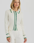 A woman stands against a plain light background, wearing the One Grey Day Royale Button Up—a white ribbed polo shirt with green trim—paired with white shorts.