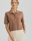 A woman in the One Grey Day Summer Cashmere Polo, a light brown short-sleeve top with buttons, and high-waisted cream pants stands against a plain light background.