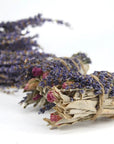The Faire Lavender, Sage and Rose Smudge features dried sage, lavender, and small red rosebuds tied with twine—ideal for spiritual cleansing as a smudge stick—displayed on a white surface with another bundle softly blurred behind.