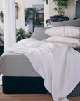 A neatly made bed features Matteo's Antik Matelasse Coverlet in white, paired with light gray sheets and white pillows. Green plants accent the room, creating a cozy, natural atmosphere.