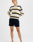 A woman stands with her hands in the pockets of dark navy shorts, wearing the Susannah Pullover by One Grey Day—a cream sweater with black and mustard stripes—paired with black flats, set against a plain white background.