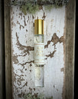 A glass "San Juan Roll On" perfume by Faire with a gold cap rests on weathered white paint and mossy wood, evoking the citrus-kissed breeze of the San Juan Islands.