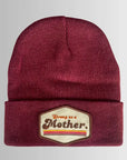 The Faire Patch Beanie Strong as a Mother is a maroon plush beanie with an adjustable cuff and a beige patch on the front that reads "Strong as a Mother" in red and black, accented by red stripes.