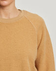 A close-up shows a person wearing the Perfectwhitetee Ziggy Reverse LS Sweatshirt in textured tan cotton fleece with a crew neckline, photographed from the shoulders up against a plain light background. Only the lower half of their face is visible.