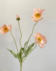 The 22'' Single Flocked Floral Poppy Spray by Faire features delicate pink silk flowers with yellow centers and green stems, including an artificial poppy and unopened bud, arranged against a light gray background.