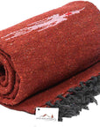 A rolled-up Rust Red Baja Thunderbird Mexican Blanket by Faire, featuring white and red patterns, black fringed edges, and a small tag with a mountain logo and "Open Road Goods." Handmade in Mexico.