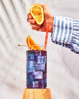 A hand in a blue and white striped shirt squeezes a lemon half over a tall glass of Faire Blackberry Jasmine Iced Tea with ice and orange slices, set on a wooden pedestal against a textured white background.