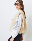 A woman with long brown hair wears large square sunglasses, the SUNCOO Diego Vest in cream recycled polyester, an embroidered-sleeve white blouse, and dark blue jeans, standing against a plain white background.