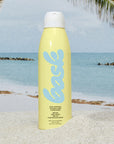 A yellow bottle of Faire Bask Mist Sunscreen SPF 30 stands upright in the sand on a beach, offering reef-safe UV protection with palm trees, ocean waves, and a blue sky in the background. Perfect for sunny days.