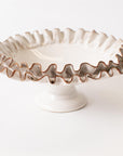 The Compote with Ruffles by Faire features a white ceramic design with a ruffled edge, short pedestal base, and brown-accented rim for rustic charm. Handcrafted home decor shown on a plain white background.