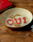 Gold heart locket necklace in a round enamel tray by Bungalow Furniture, displayed on a wooden table with books nearby.