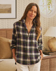 A woman with long brown hair wears the Frank & Eileen Eileen Button Up Shirt F25 and white pants, smiling indoors with hands in her pockets. The room features patterned wallpaper, a lamp, framed art, and neutral-toned decor.
