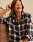 A woman with long brown hair smiles indoors, wearing the Frank & Eileen Eileen Button Up Shirt F25—a relaxed-fit plaid shirt in black, white, yellow, and red. She touches her hair and channels California style: confident, comfortable, and happy.