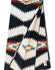 The Tsachila Blanket by Faire is a folded recycled acrylic throw with bold black, white, brown, blue, and red zigzag and diamond patterns—ideal for boho decor. A fabric tag is visible on one corner.