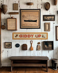 A rustic white wall displays vintage decor—frames, maps, paddles, fishing gear, and the Faire "Giddy Up" Felt Banner—above a wooden bench on polished floors. Sunlight enhances the space’s cozy, nostalgic charm.