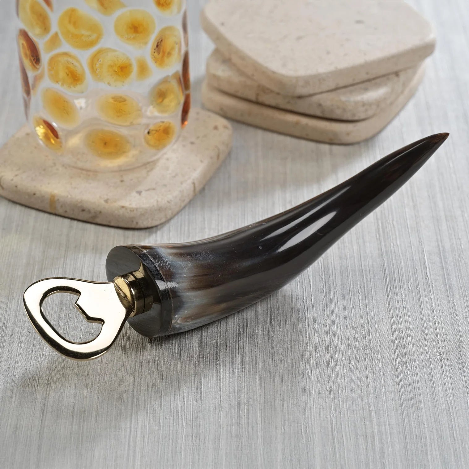 The Zodax Horn Bottle Opener features a dark, polished horn handle and metal tip, shown on a light wood surface beside a textured glass and stacked stone coasters—distinctive rustic barware for any setting.