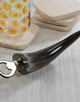 The Zodax Horn Bottle Opener features a dark, polished horn handle and metal tip, shown on a light wood surface beside a textured glass and stacked stone coasters—distinctive rustic barware for any setting.