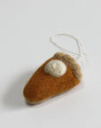 The Faire Felted Ornament is a handmade pumpkin pie slice crafted from wool, featuring a dollop of white cream and an attached string loop, displayed on a white surface.
