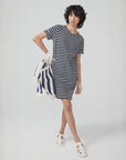 A person with short curly hair stands against a plain light background, wearing a black and white striped dress and white sandals, holding Kule’s The All Over Striped Tote.