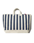 The All Over Striped Tote by Kule is a large cotton canvas bag with vertical navy blue and white stripes, two short handles, and a small red and blue tag at the top right corner. Photographed against a white background.