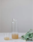 The Creative Co-op Decanter With Glass, partially filled with a light brown liquid, rests on a white surface beside a folded green fringed cloth—elegant tableware for any setting against a plain light gray background.