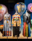 Three mannequins display “Paris In Fashion” by Simon & Schuster Inc., wearing vibrant, artistic outfits inspired by Paris, in front of painted hot air balloons and a dark backdrop in a stylish window display.