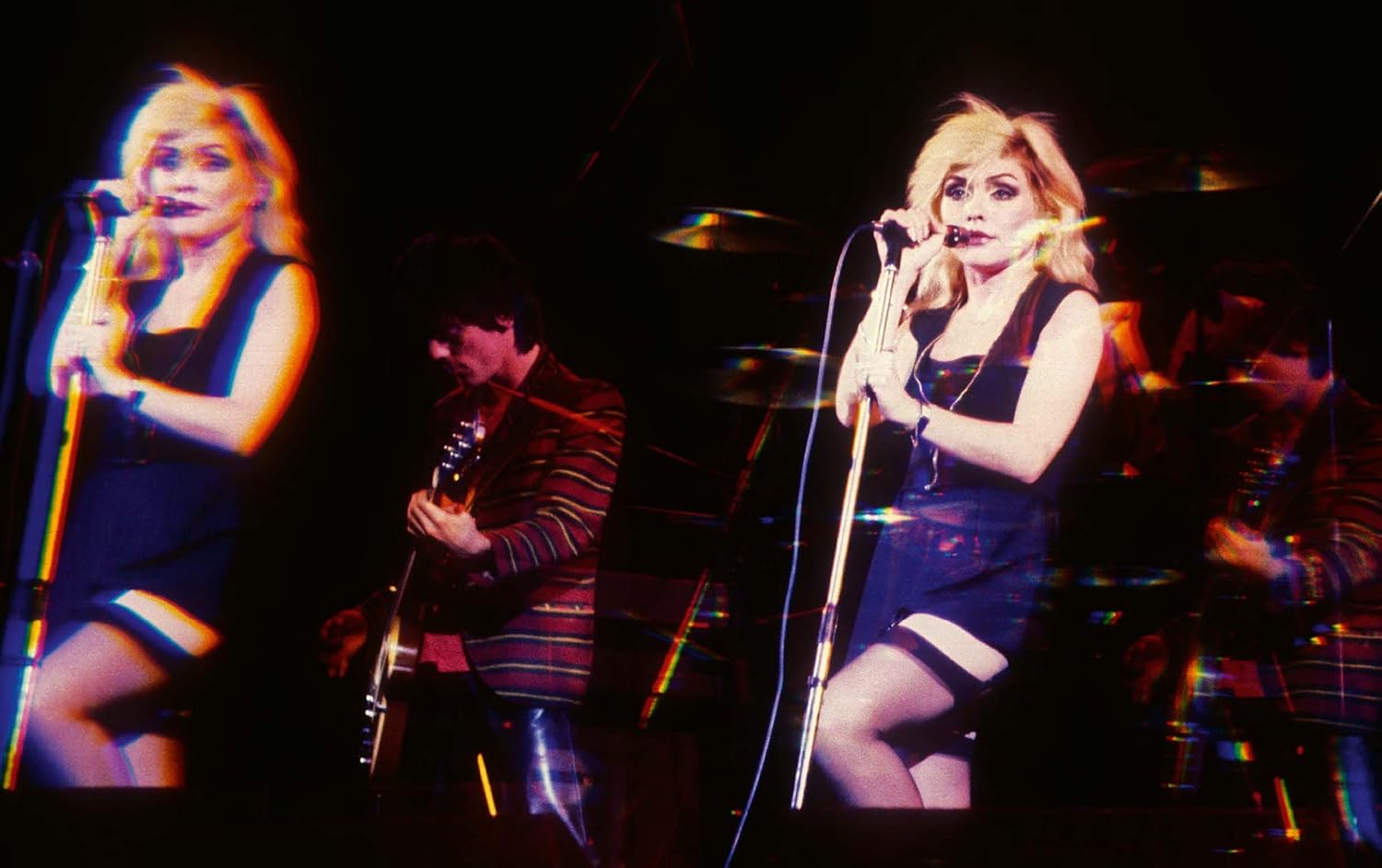 A prismatic, double-exposure photo with vivid colors from "Blondie In Camera 1978" by Simon & Schuster Inc. shows a blonde woman singing on stage beside a guitarist, echoing classic Blondie images with motion blur effects.