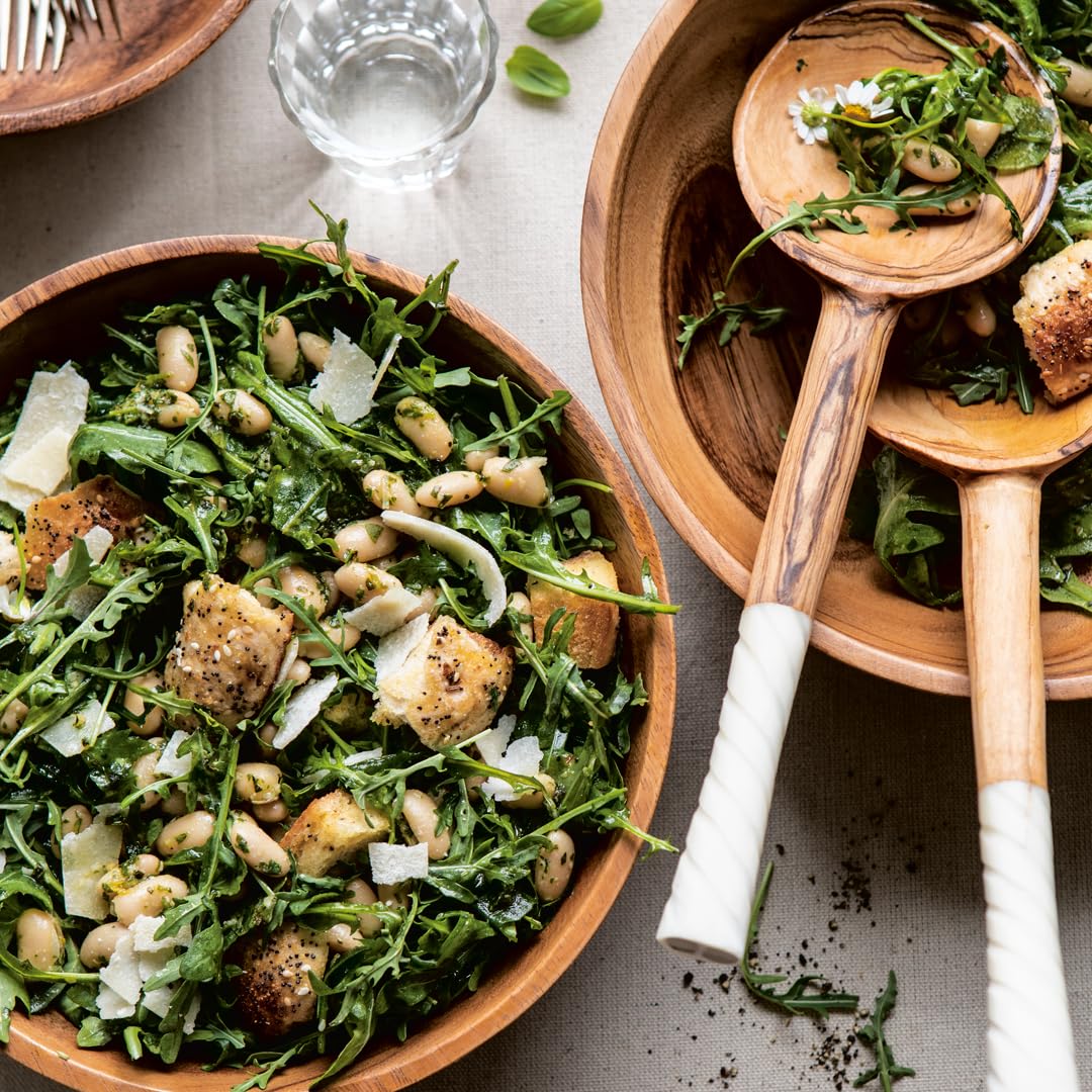 Inspired by Half Baked Harvest Super Simple from Random House, two wooden bowls of arugula salad with white beans, croutons, and parmesan are served with utensils on a light tablecloth, alongside water and scattered greens.