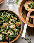 Inspired by Half Baked Harvest Super Simple from Random House, two wooden bowls of arugula salad with white beans, croutons, and parmesan are served with utensils on a light tablecloth, alongside water and scattered greens.