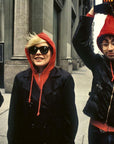 Three people walk on a city sidewalk. One, in sunglasses and a red hoodie under a black jacket, channels Blondie’s style as seen in Martyn Goddard's photos from "Blondie In Camera 1978" by Simon & Schuster Inc., with urban buildings behind them.