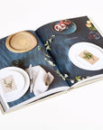 The book "Sunday Suppers" by Random House is open to a scene of two white plates, beige napkins, greenery on a dark blue cloth, and a wooden bowl with fruit—an inviting table set for communal cooking and gathering friends.