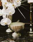 The Accent Decor Lita Compote L in antiqued silver rests on a dark marble surface beside a tall vase of white orchids and roses, with a gold candlestick positioned in the background.