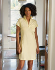 A woman with curly hair wears the Frank & Eileen Lauren Polo Dress in pale yellow, standing in a bright hallway with a table and large window, showcasing a preppy classic style.