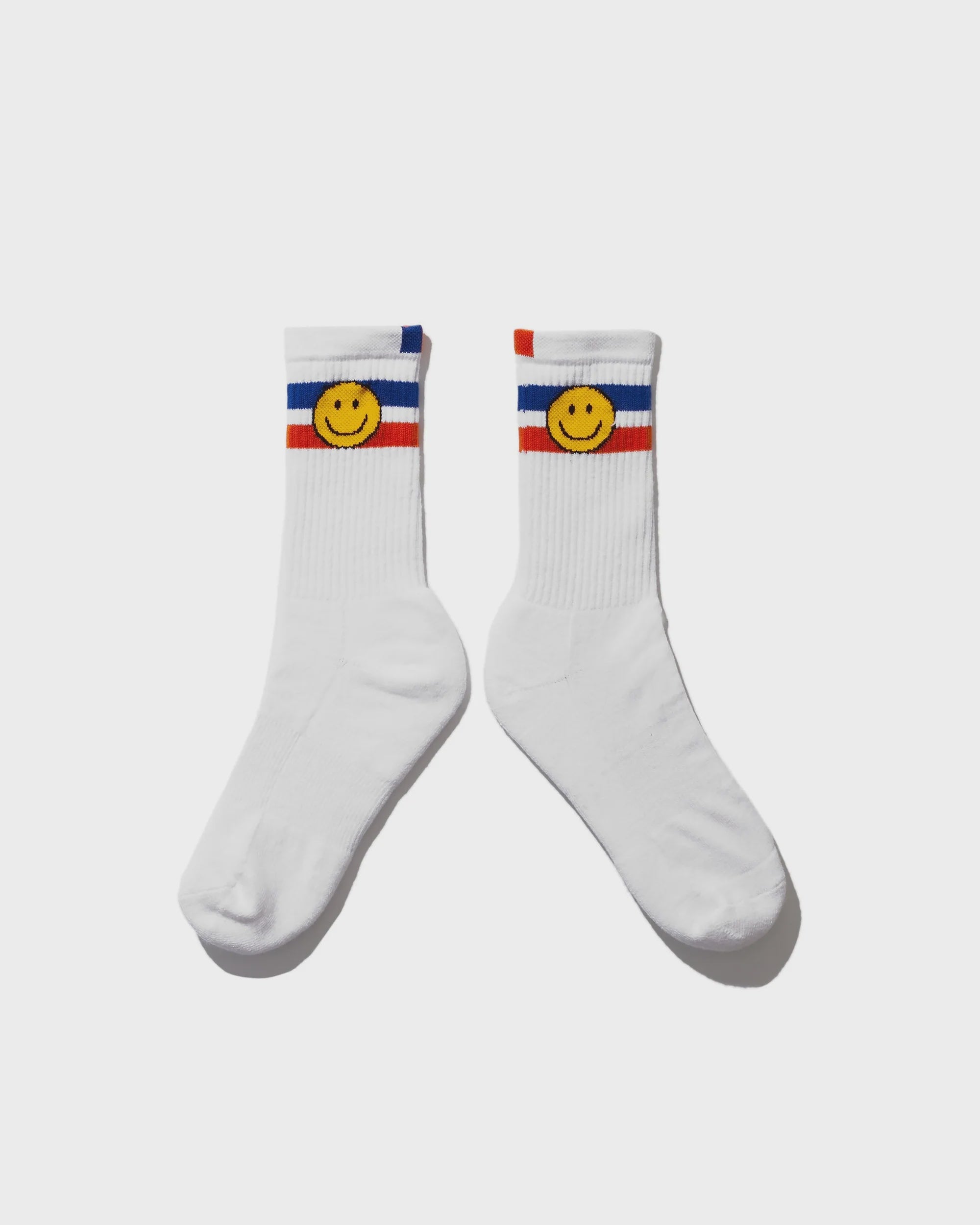 Kule's The Women's Ribbed Smile Sock WHT are white crew socks with blue and red stripes near the top and a yellow smiley face on each sock; they’re ribbed and machine washable for easy care.