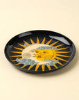The Indaba Sun and Moon Tray is a round, black ceramic piece with enchanting sun and moon faces, golden rays, and blue clouds on a light beige background—ideal for whimsical accent decor.
