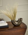 The Franz Vase by Accent Decor, crafted from recycled cast aluminum with a dark oxidized finish and filled with dried pampas grass, sits on a dark wooden table beside a stack of books against a light-colored wall.