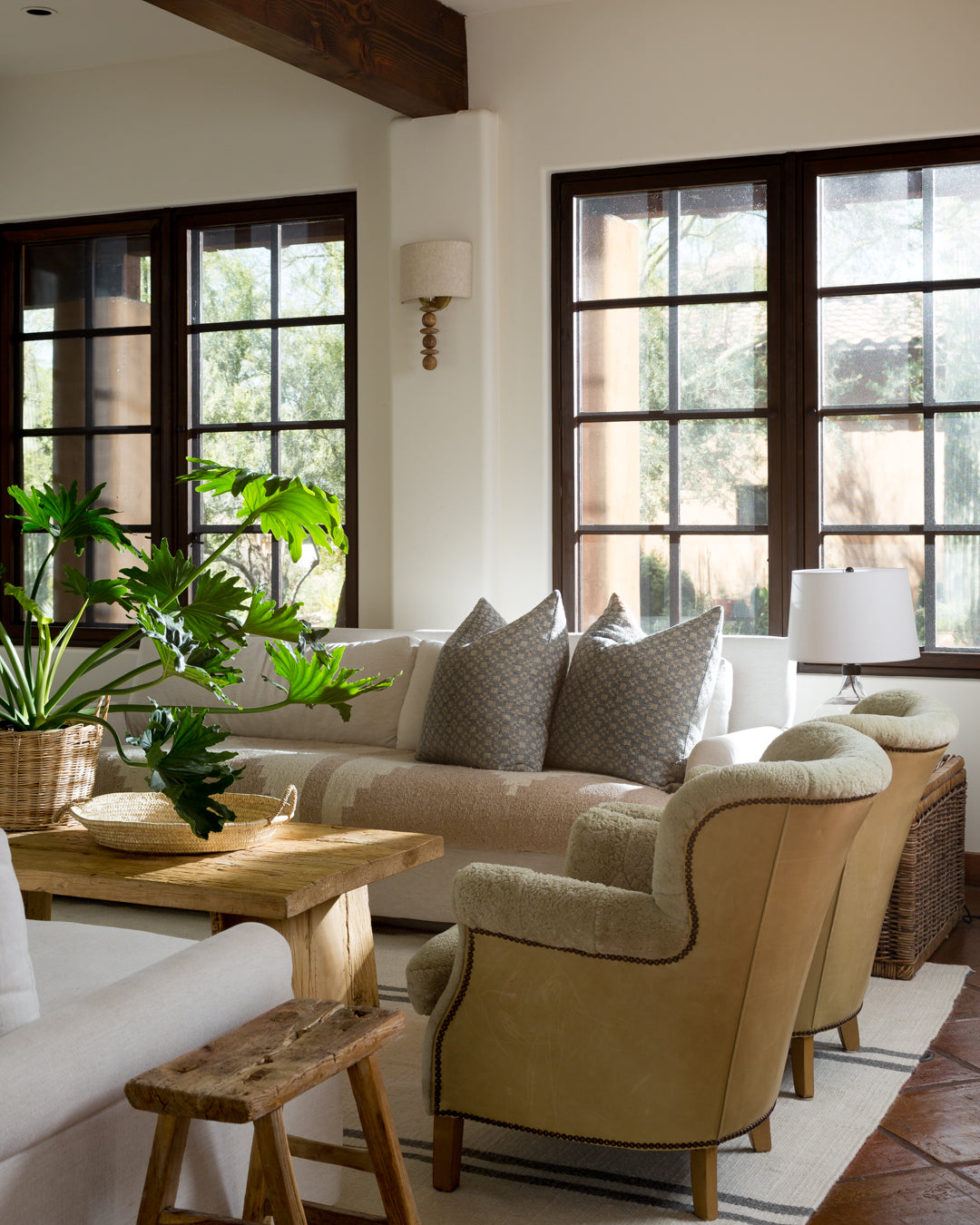 A cozy living room with large windows, beige sofas, a wicker side table, and wooden accents. Natural light illuminates the space decorated with plants and soft pillows.