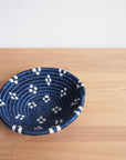 The Kabaya Small Bowl by Amsha - Faire, a round, shallow Rwanda basket with a blue and white geometric pattern, rests on a light wooden surface against a white textured wall.
