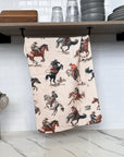 The Faire Rodeo Towel, featuring a cowboy-on-horseback pattern, hangs from a shelf below stacked dishes and glasses, above a countertop with a container of assorted kitchen utensils.