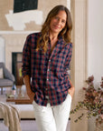 A woman with long brown hair stands indoors, smiling softly in the Frank & Eileen Eileen Button Up Shirt F25— a relaxed-fit red and navy plaid shirt with flattering button placement—paired with white pants, leaning against a beige wall.