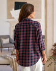 A woman with long brown hair stands indoors, facing away, wearing the Frank & Eileen Eileen Button Up Shirt F25 in red, blue, and white plaid with rolled-up sleeves and white pants. The room has neutral decor and artwork in the background.