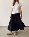 A woman stands indoors against a light wall, wearing Hey Gang’s The Ranch Skirt—a long black cotton skirt with an elastic waist—paired with a white short-sleeve t-shirt and beige clogs. Her face is not visible.